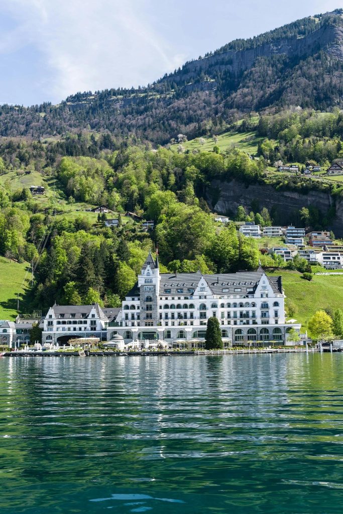 A grand white lakeside hotel at the base of a lush green mountain reflected in calm green water.