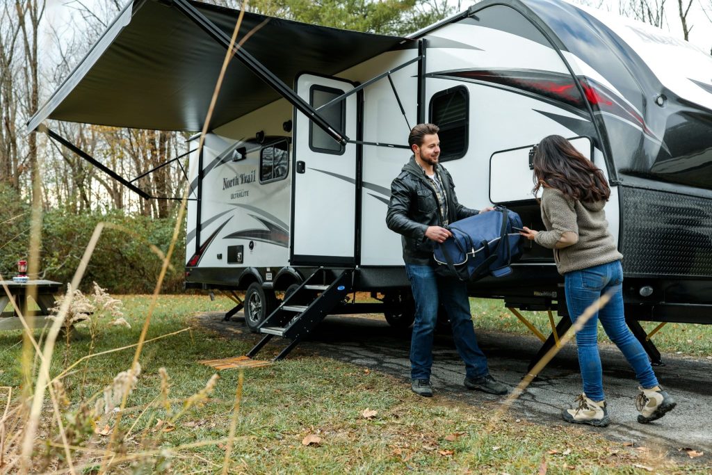 A couple loading a blue duffel bag into the storage compartment of a North Trail travel trailer at a campsite.