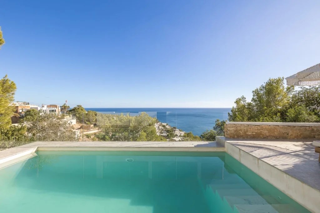 An elevated turquoise pool with a glass railing looks out over a green hillside and the blue Mediterranean Sea.