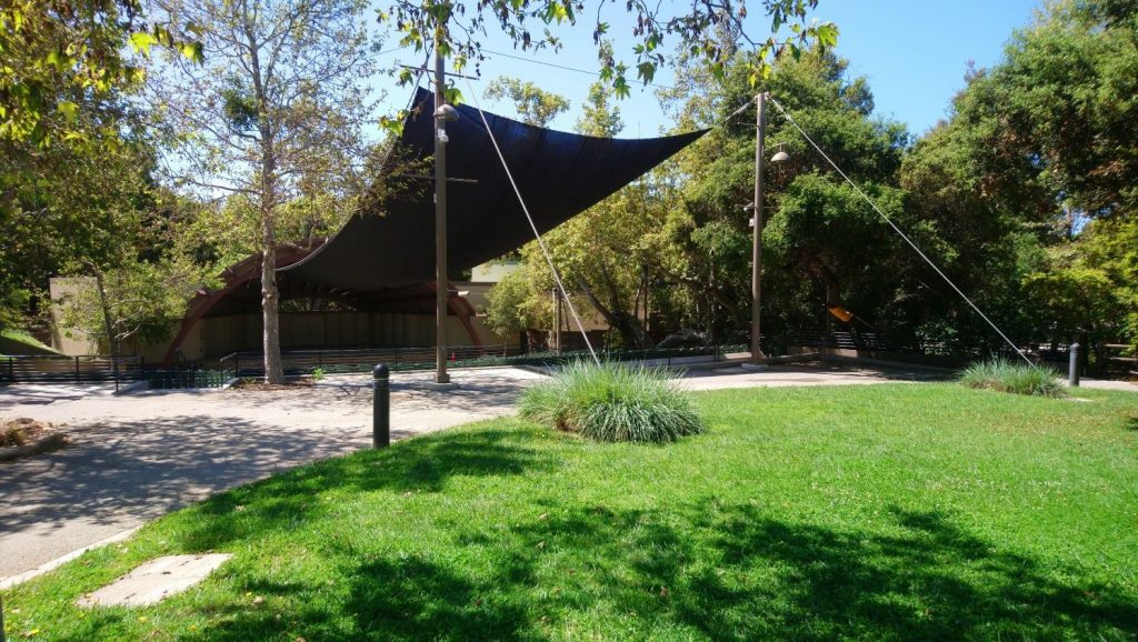 Outdoor amphitheater with a dark shade sail and wooden stage surrounded by green trees and a sunny lawn in Ojai Music Festival