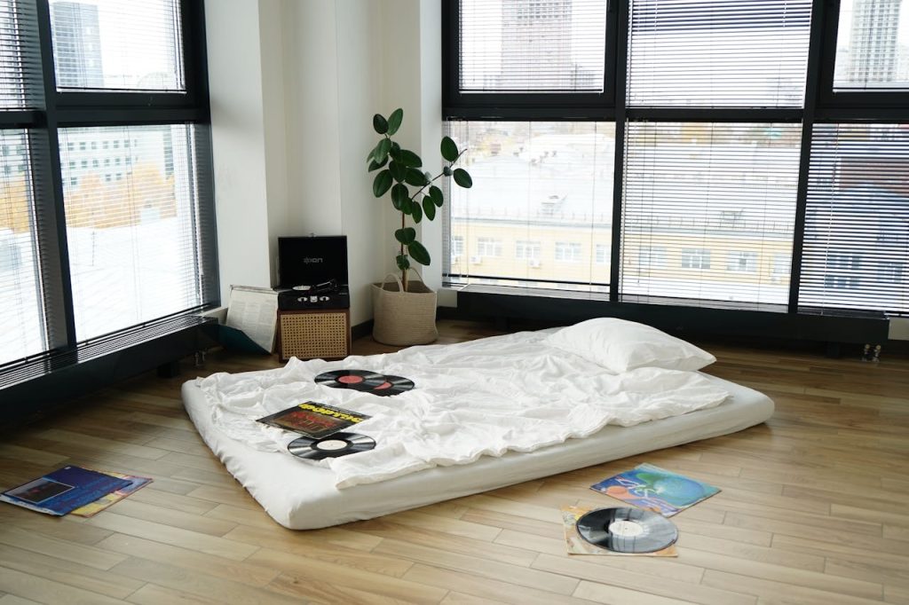 A minimalist room with a mattress on the floor, scattered vinyl records, a record player, and a large window.