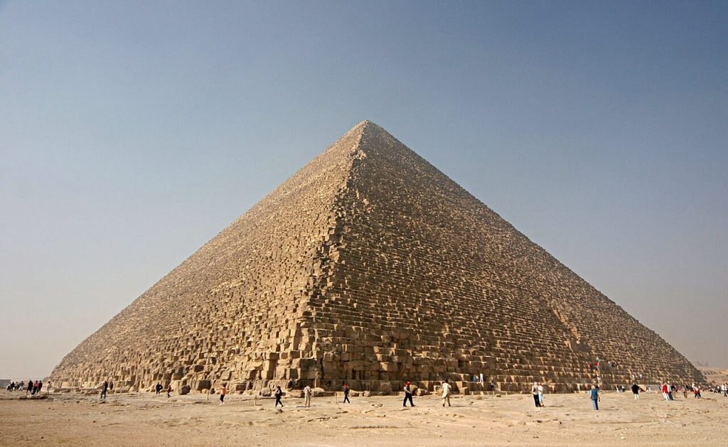 The Great Pyramid of Giza under a clear sky with small figures of tourists walking along the sandy base for scale.