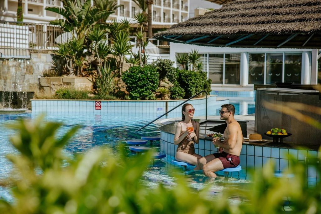 A couple relaxes on underwater stools at a resort swim-up pool bar, drinking cocktails on a sunny day.