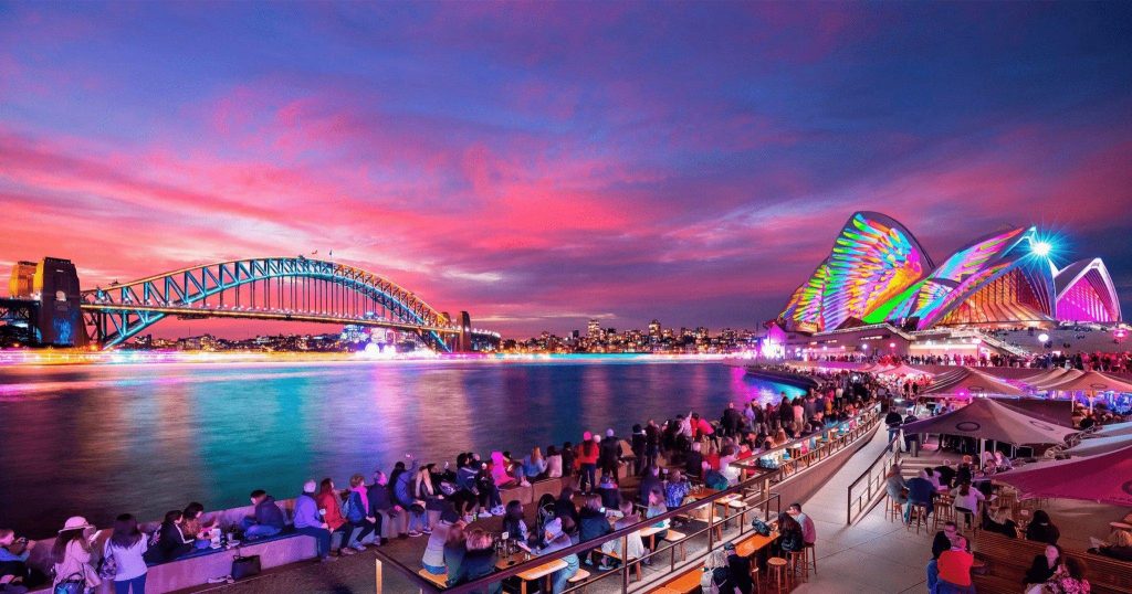Crowds watch Vivid Sydney light projections on the Sydney Opera House and illuminated Harbour Bridge at twilight.