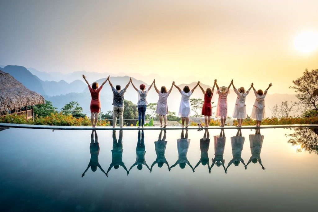 A group of friends hold hands with raised arms at an infinity pool, reflecting perfectly against a mountain sunrise.