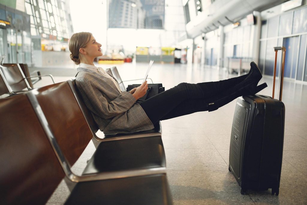 A relaxed woman in a plaid blazer rests her feet on her black suitcase while waiting in an airport terminal.