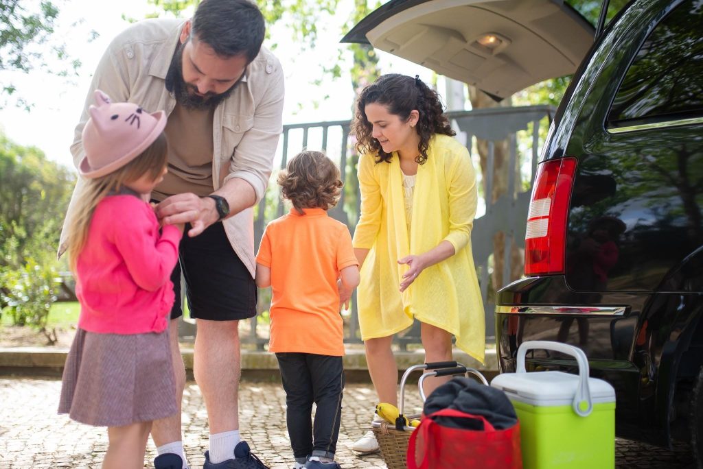 A family packs a picnic basket and cooler into the back of a black car for a weekend road trip.