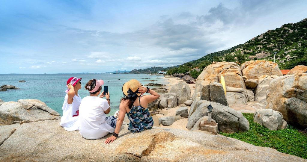 Three women sitting on coastal rocks overlooking a tropical beach and turquoise ocean on a sunny day.