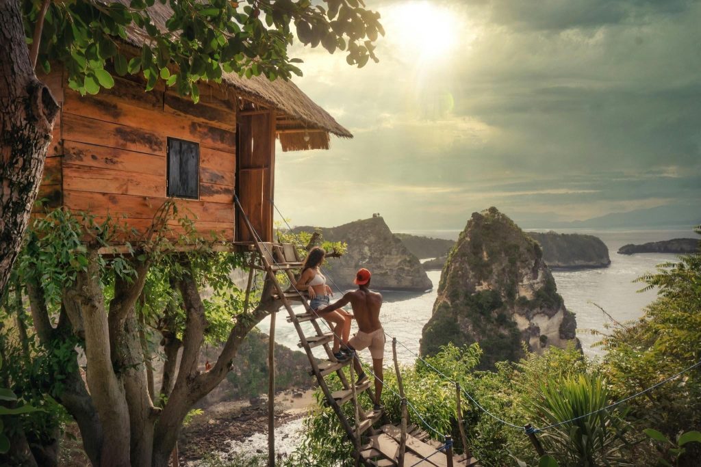 A couple on a wooden treehouse ladder overlooking limestone cliffs and the ocean in Nusa Penida, Bali at sunset.