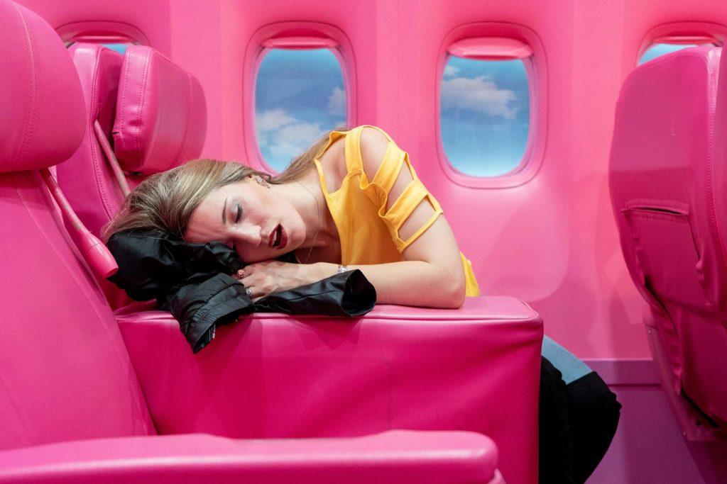 A woman in a yellow top sleeping on a black jacket inside a vibrant, all-pink airplane cabin with blue sky windows.