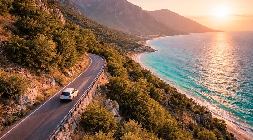 A white car drives along a winding coastal road overlooking a turquoise beach and mountains at sunset.