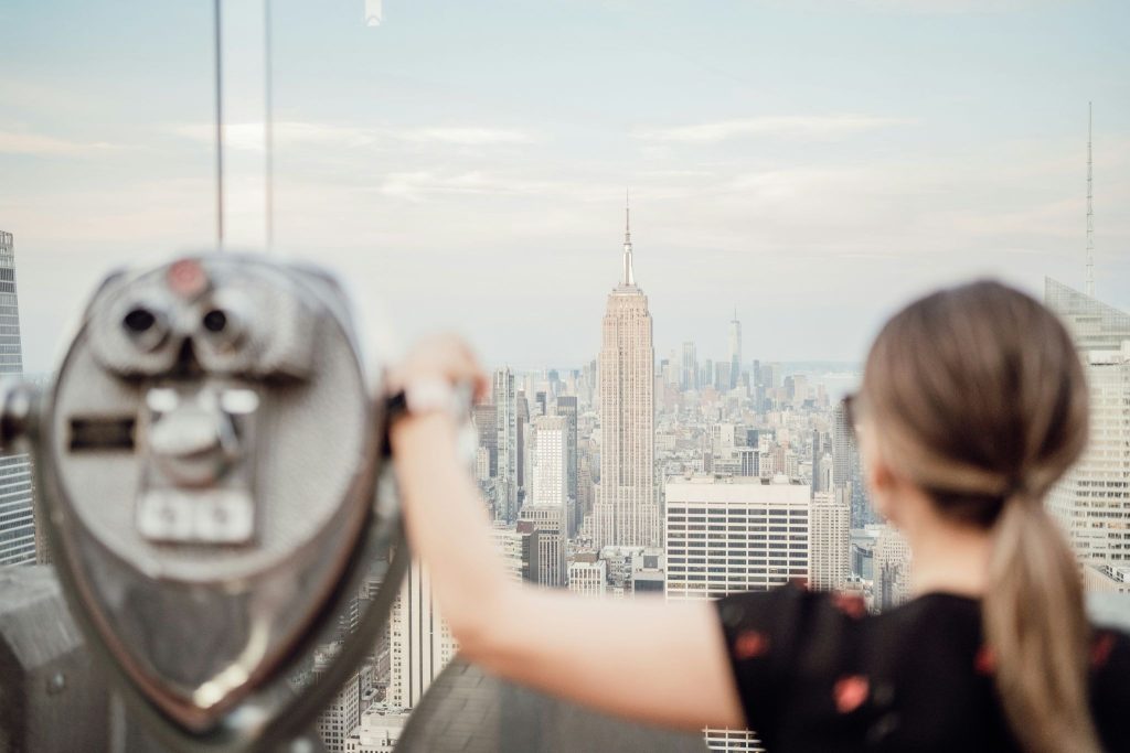 Blurred woman and observation deck binoculars framing a sharply focused, bright view of the Empire State Building and NYC skyline.