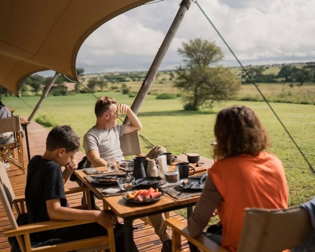 Family enjoying breakfast on a wooden deck under a luxury safari tent, looking out over a lush green savanna landscape.