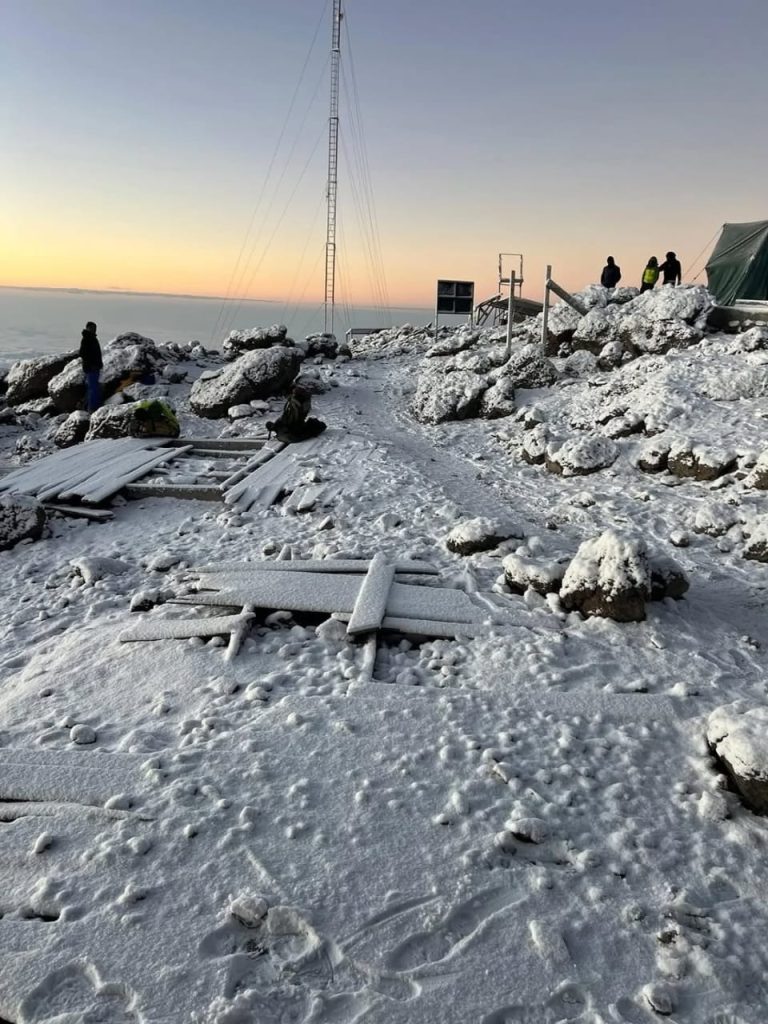 Snowy mountain summit camp above the clouds at sunrise, featuring hikers, a green tent, wooden planks, and a tall mast.