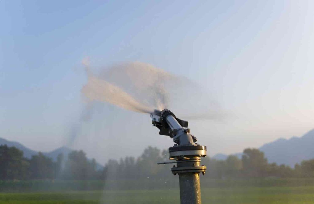 An agricultural water cannon sprinkler spraying a powerful mist over a green farm field at sunrise.