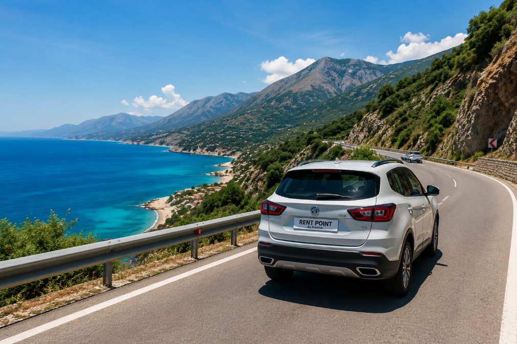 A white rental SUV drives along a winding coastal highway overlooking the turquoise sea and mountains in Albania.