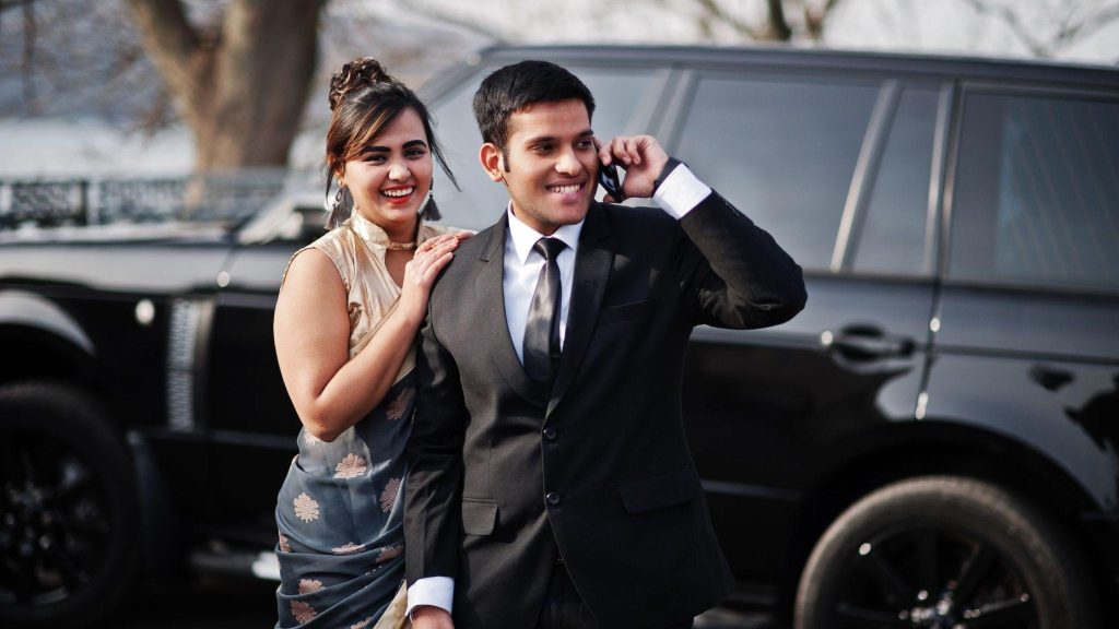 Elegant South Asian couple by a black luxury SUV; the man in a suit talks on a smartphone while the woman in a saree smiles.