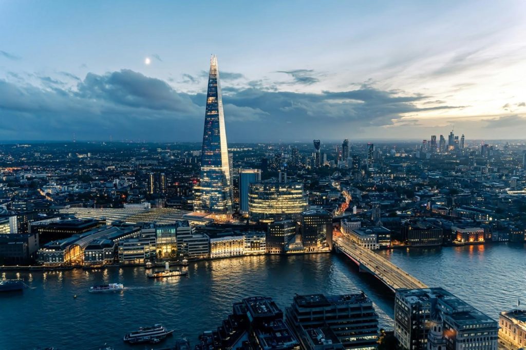 Aerial view of the illuminated Shard skyscraper towering over the River Thames and the sprawling London city skyline at twilight.