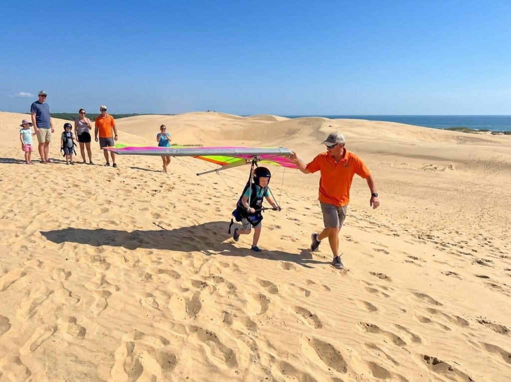Young boy learning to hang glide on large sand dunes, running with a colorful glider alongside his instructor under a blue sky.
