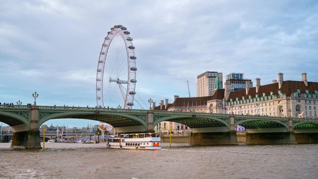 White passenger boat on the River Thames passing under Westminster Bridge, with the iconic London Eye in the background.