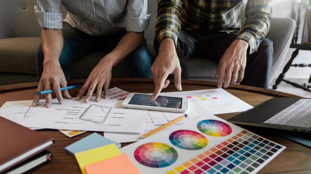 Two designers collaborating at a wooden table, pointing at a digital tablet surrounded by UI wireframes and color palettes.