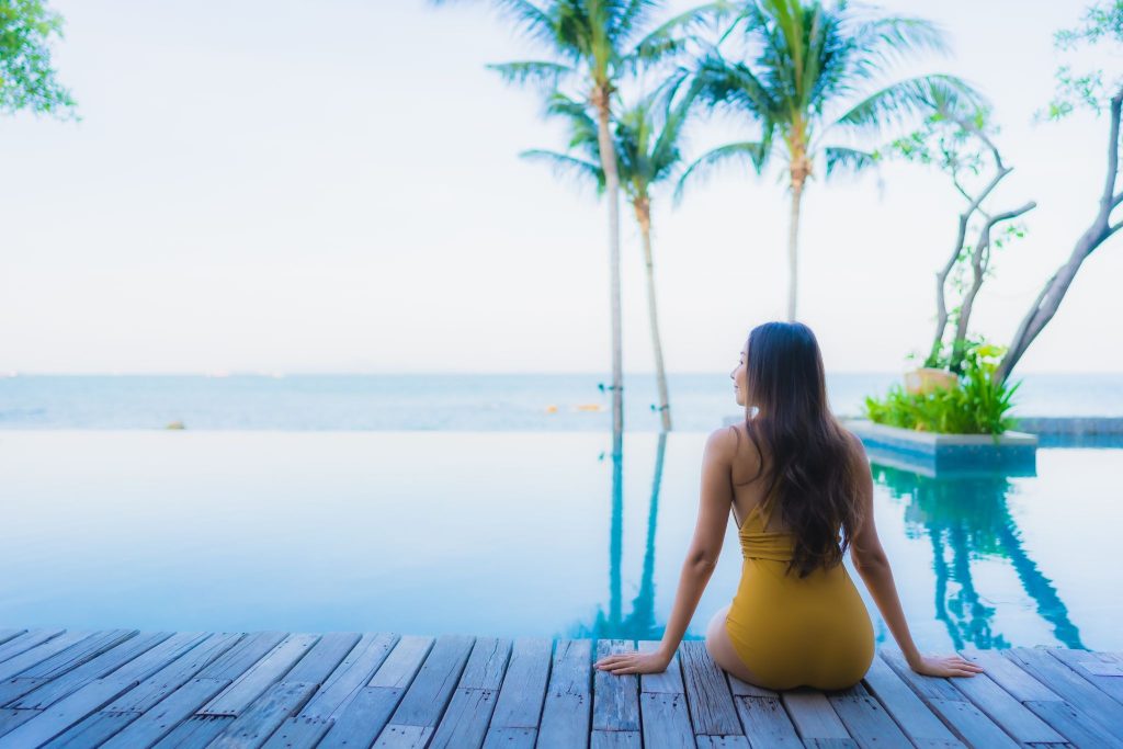 Woman in a yellow swimsuit relaxing on a wooden deck by a tropical infinity pool overlooking the ocean.