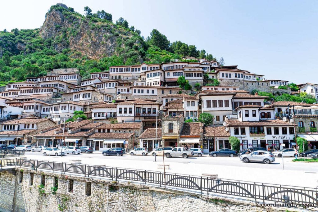 Historic white Ottoman-style houses with prominent windows stacked on a green hillside in Berat, Albania, under a blue sky.