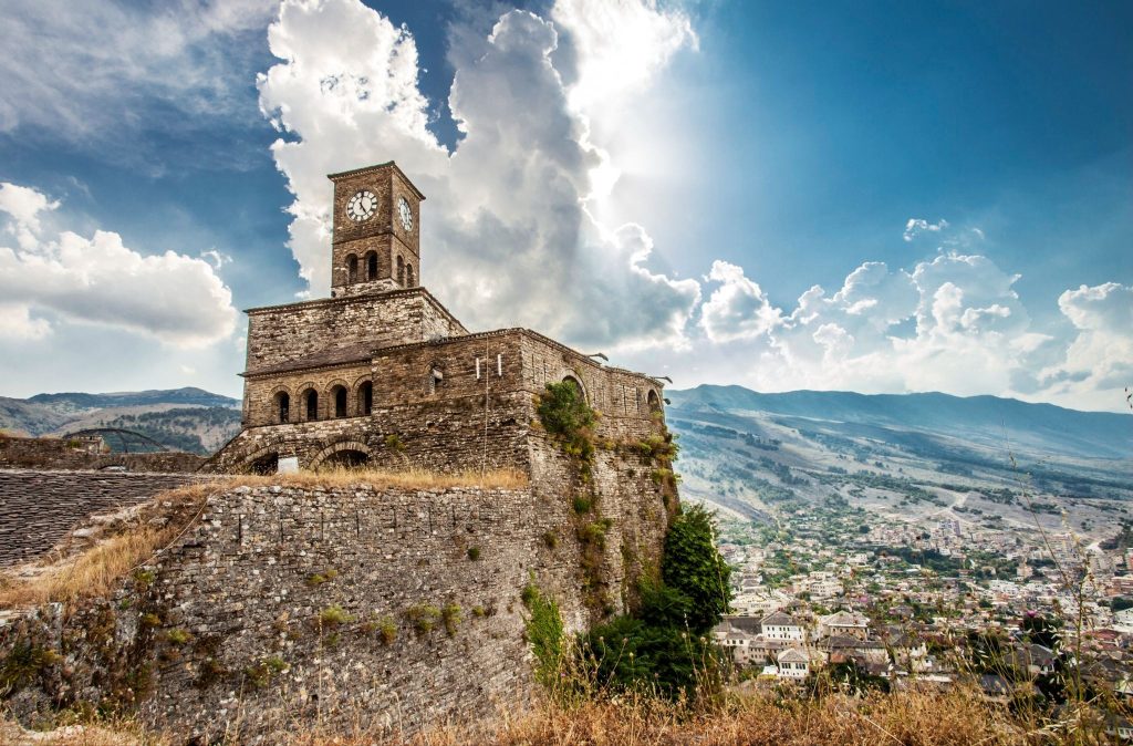 Historic Gjirokastër Fortress and clock tower on a cliff overlooking a sprawling Albanian valley town under a dramatic sky.
