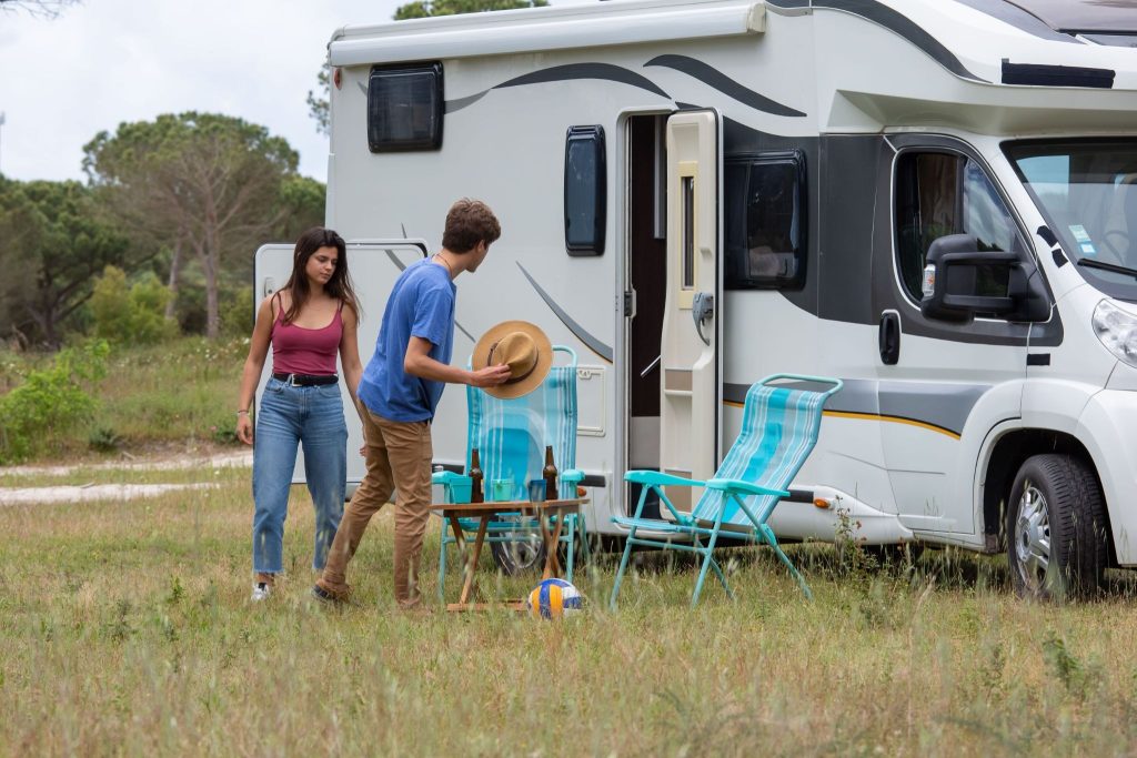 A young couple sets up striped lawn chairs and a small table next to their white motorhome at a grassy campsite.