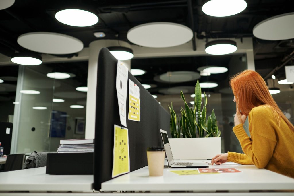 Focused red-haired woman in a yellow sweater working on a laptop at a modern white office desk with an acoustic divider.