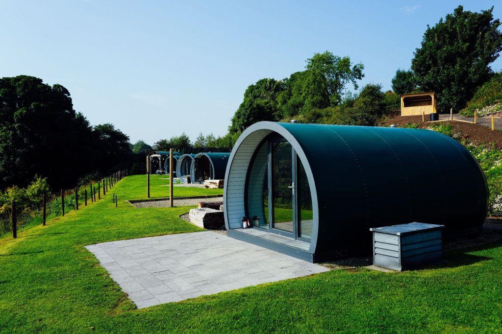 Row of dark green modern glamping pods with glass fronts on a sunny, lush green hillside under a clear blue sky.