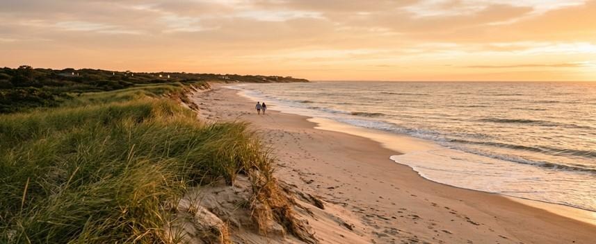 A distant couple walks along a wide sandy beach bordered by grassy dunes during a radiant golden sunset.