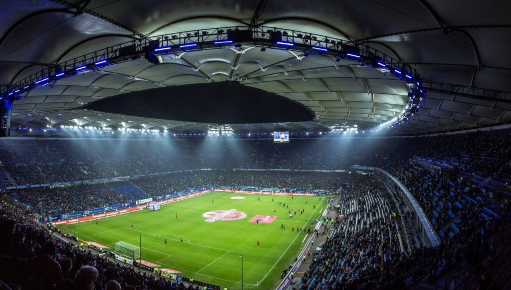 High-angle view of an illuminated football stadium with a green pitch and crowds under a blue-lit domed roof.