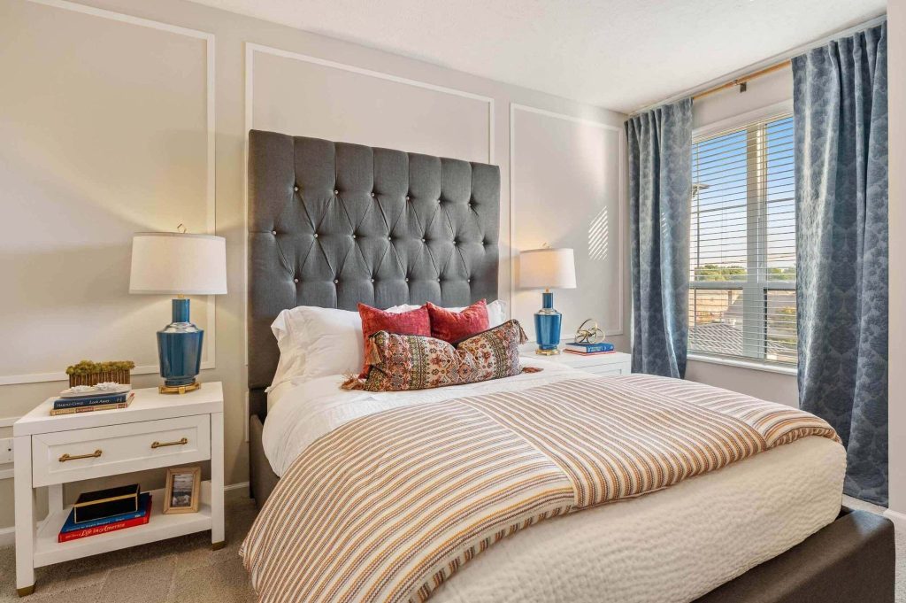 Bright contemporary bedroom with a tall grey tufted headboard, blue table lamps, and white nightstands near a sunny window.
