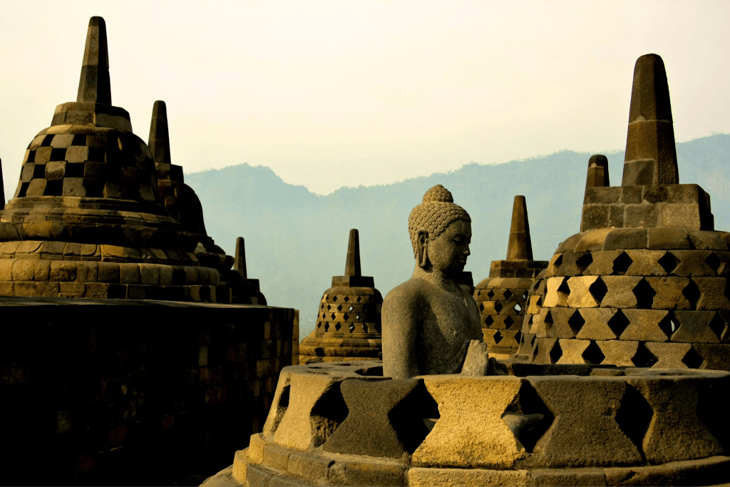 Stone Buddha statue in a meditative pose inside a ruined stupa at Borobudur Temple, with intact stupas and hazy mountains.