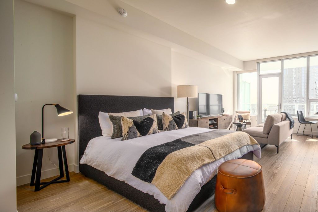 Modern open-plan bedroom with textured bedding, leather ottoman, and a cozy living space facing large, bright city windows.