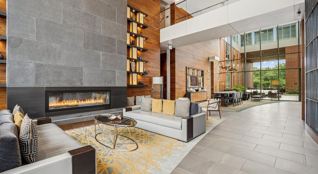 Modern lobby lounge with a grey stone fireplace, sleek facing sofas with yellow pillows, wood paneling, and large glass windows.