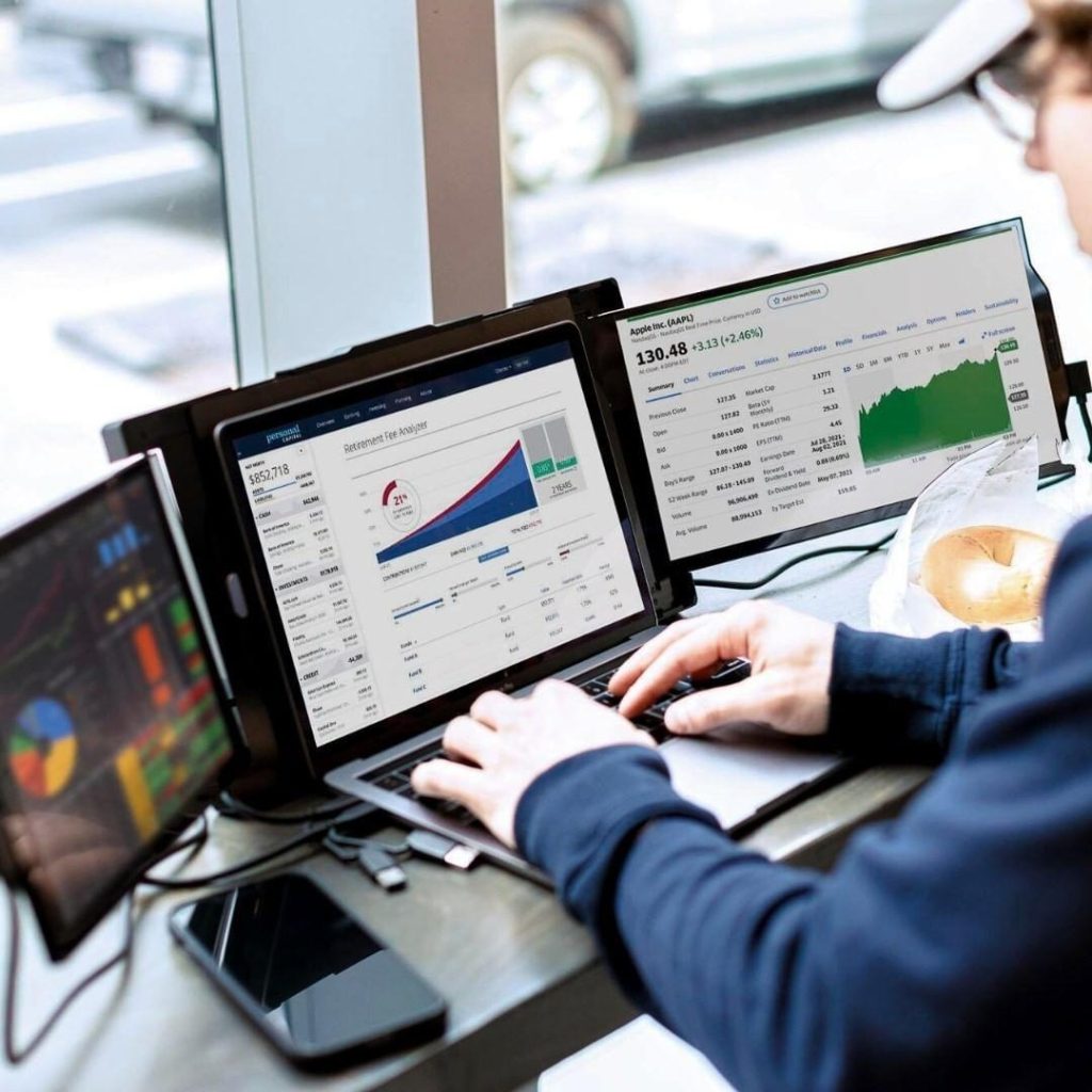 Person typing on a tri-screen laptop setup displaying financial dashboards and stock market charts near a bright window.