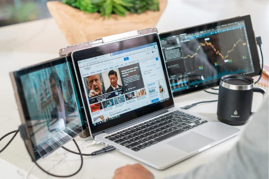 Three-screen laptop setup on a marble desk displaying financial news, a video stream, and stock market trading charts.