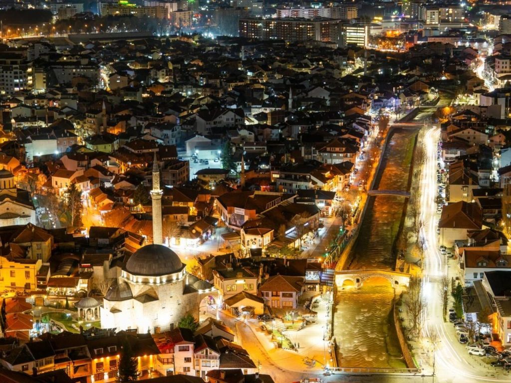 Elevated night view of a historic city center featuring a domed mosque, an arched stone bridge over a river, and traffic trails.