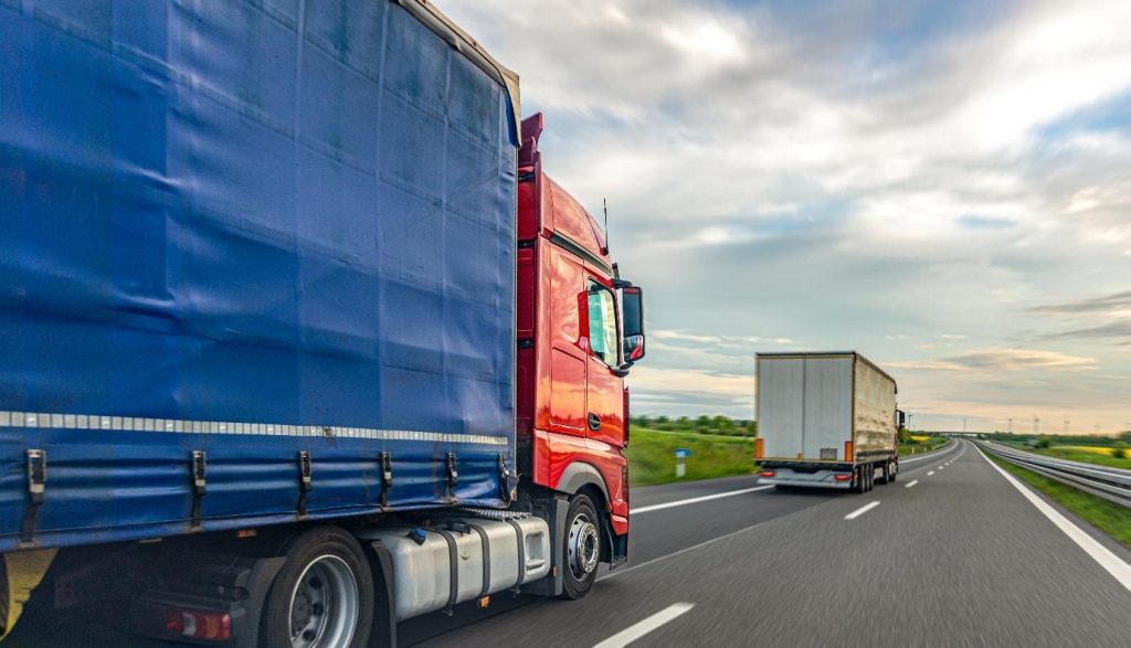 Red semi-truck with a blue curtain trailer driving quickly on a highway, with road motion blur and a cloudy sky above.