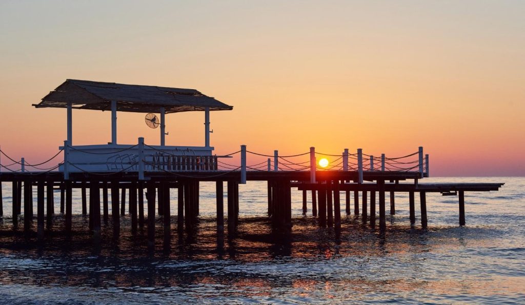 Silhouette of a wooden pier and pavilion extending into a calm ocean during a vibrant orange and pink sunset.