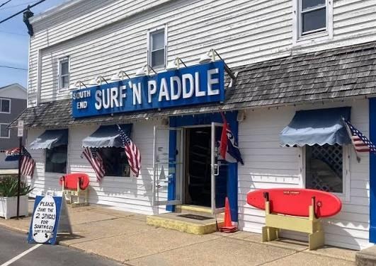 Exterior of South End Surf 'n Paddle shop featuring a blue sign, American flags, and a red paddleboard on the sidewalk.