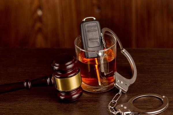 Car keys submerged in a glass of liquor, surrounded by a judge's gavel and metal handcuffs on a dark wooden table.