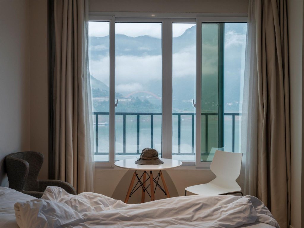 Hotel room seating area by a large window overlooking a serene river, misty green mountains, and a distant red bridge.