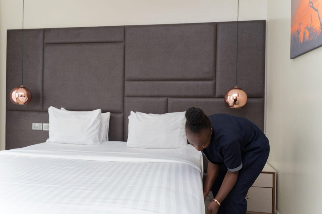 Hotel housekeeper in a blue uniform making a crisp white bed beneath a modern brown geometric headboard and copper lights.