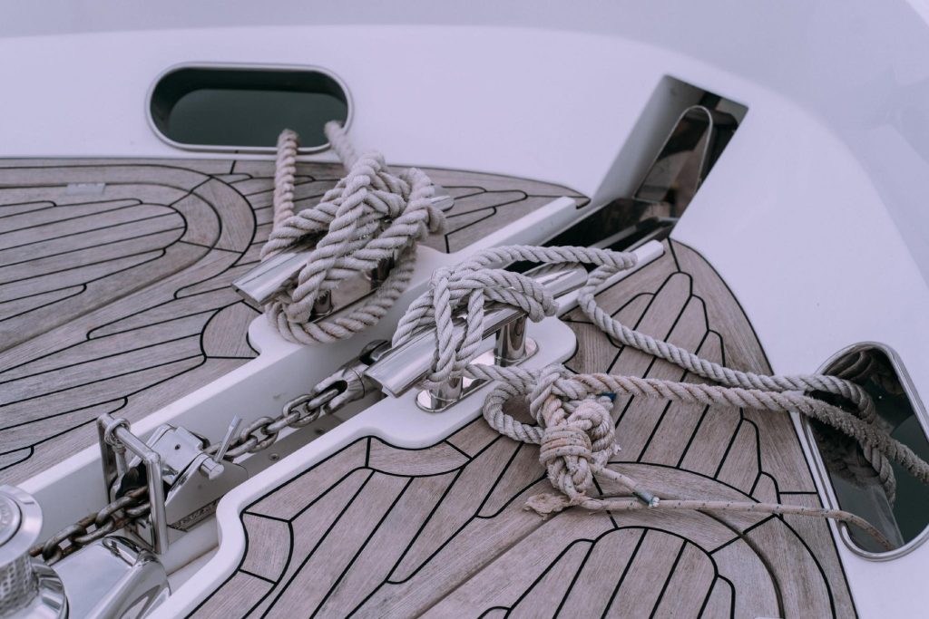 Polished stainless steel cleats tied with white marine rope and an anchor chain on the teak deck of a luxury yacht's bow.