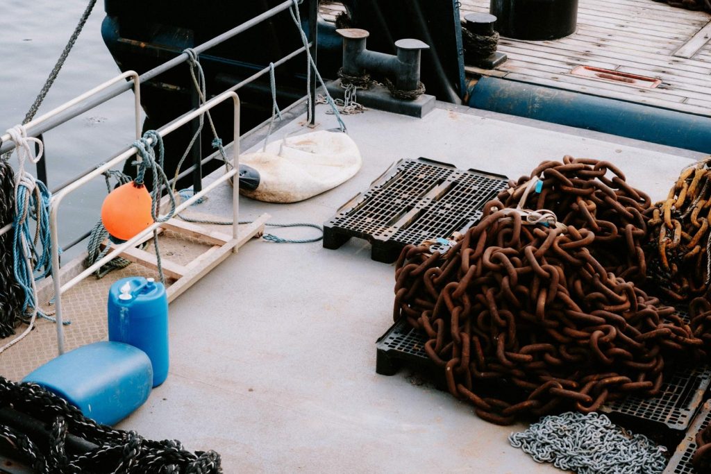 Heavily rusted maritime chains piled on pallets on a vessel deck, next to a bright orange buoy and blue plastic barrels.