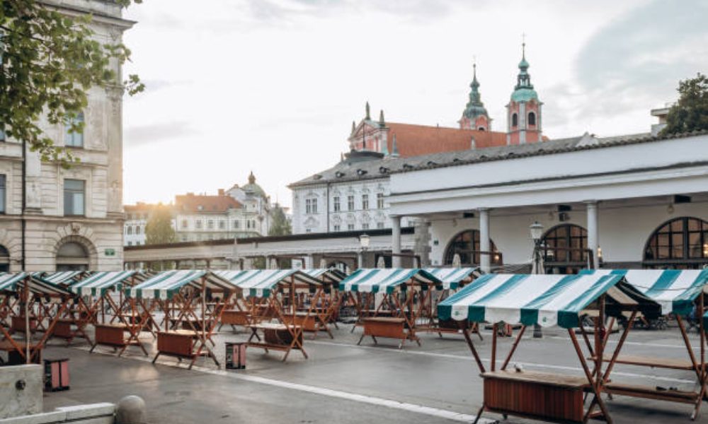 Empty wooden stalls with green and white striped awnings in Ljubljana Central Market, backed by historic city architecture.