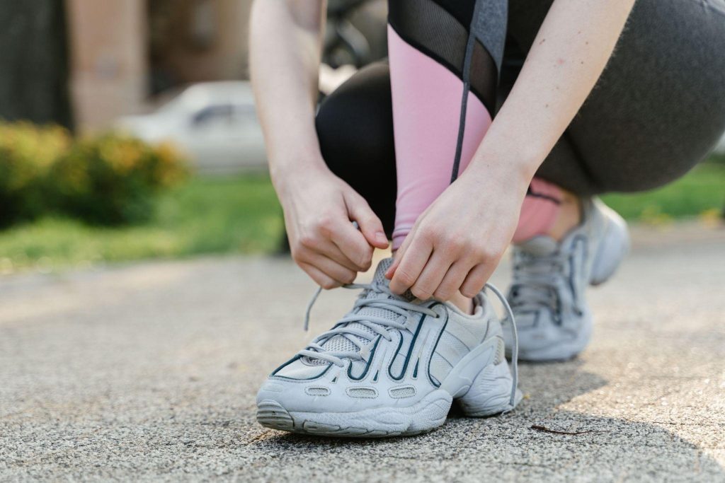 Person crouching on an outdoor paved path tying the laces of their light grey chunky running shoes before a workout.
